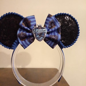 Puede incluir: Una diadema con dos orejas circulares cubiertas de lentejuelas negras y ribete de pompones azules. Un lazo central muestra rayas azules y el escudo de Ravenclaw con un águila y el texto Ravenclaw impreso en blanco y negro.