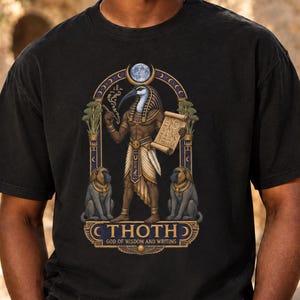 May include: A black short-sleeve t-shirt featuring a graphic of the Egyptian deity Thoth with an ibis head, holding a scroll and a stylus. The design includes two baboons, a moon, and the text THOTH GOD OF WISDOM AND WRITING.