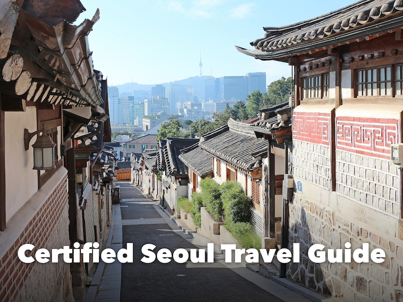 May include: A narrow street in Seoul lined with traditional Korean hanok houses featuring tiled roofs and stone walls, with a modern city skyline and N Seoul Tower visible in the distance. The text Certified Seoul Travel Guide is overlaid.