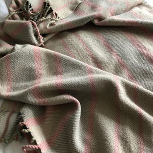 Pode incluir: Cachecol xadrez bege e rosa com franjas. O cachecol tem uma textura tecida com listras rosa verticais sobre um fundo bege. As bordas são finalizadas com franjas em uma paleta de cores correspondente. O cachecol está sobre uma superfície branca.