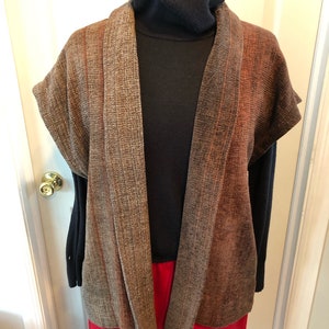 May include: A brown and orange sleeveless cardigan with a shawl collar. The cardigan is made of a textured fabric and has a relaxed fit.