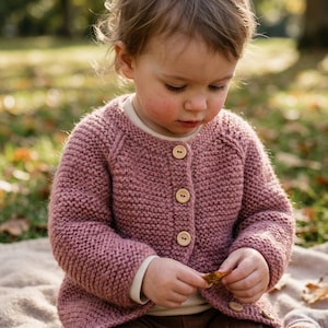 May include: A dusty rose-colored, long-sleeved knit cardigan featuring a round neckline and a front button closure with four light-colored wooden buttons. The garment is crafted with a textured garter stitch pattern and has raglan sleeves.