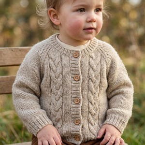 May include: A beige cable-knit cardigan featuring a round neckline, long sleeves, and a front button closure with five small brown buttons. The knit pattern includes vertical cable details on the front panels and ribbed cuffs.