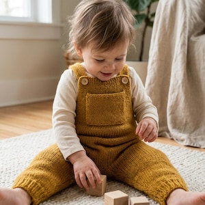 May include: A mustard yellow knitted overall featuring a front pocket and two light-colored wooden buttons at the shoulder straps. The garment is worn over a long-sleeved cream-colored shirt, showcasing a textured knit pattern throughout.