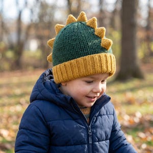 May include: A dark green knitted beanie featuring a mustard yellow ribbed cuff and a row of matching mustard yellow triangular spikes extending from the crown to the back, mimicking the appearance of a dinosaur crest.