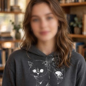May include: A charcoal gray hooded sweatshirt featuring a white graphic illustration of a cat standing on a stack of books with paw prints on the cover. Below the illustration, the text reads, I teach, my cat grades.