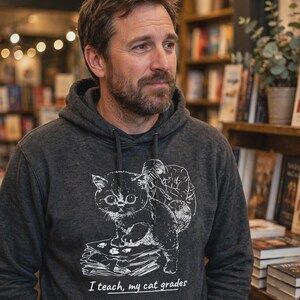 May include: A charcoal gray hooded sweatshirt featuring a white line art graphic of a cat standing on a stack of papers with a person behind it. The text below the graphic reads, "I teach, my cat grades," with a line under the word teach.