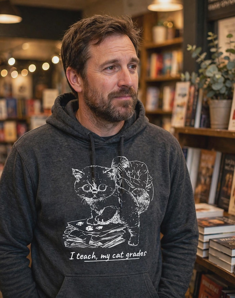 May include: A charcoal gray hooded sweatshirt featuring a white line art graphic of a cat standing on a stack of papers with a person behind it. The text below the graphic reads, "I teach, my cat grades," with a line under the word teach.