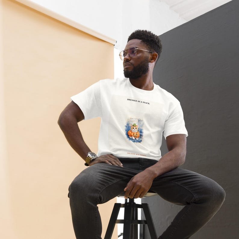 K&ouml;nnte beinhalten: Ein wei&szlig;es Kurzarm-T-Shirt mit einer Grafik einer Katze, die einen K&uuml;rbishut tr&auml;gt und in einem K&uuml;rbis sitzt. Der Text &uuml;ber der Grafik lautet DRESSED AS A SNACK und der Text unter der Grafik lautet ONE CUTE BUT SQUASHY.