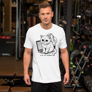 May include: A white short-sleeved t-shirt featuring a black and white graphic of a cat sitting on an open laptop with a person behind it. The text below the graphic reads $ my cat shipped it. The design is centered on the chest area.