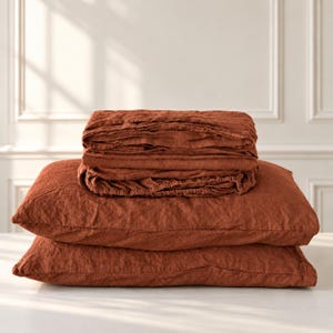 May include: A set of rust-colored bedding featuring two rectangular pillows stacked at the bottom and a folded flat sheet or duvet cover resting on top. The fabric has a visible textured weave and a matte finish in a deep terracotta tone.