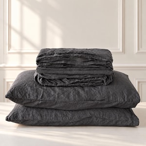 May include: A set of dark charcoal gray bedding featuring two rectangular pillows stacked horizontally, topped with a neatly folded, textured fabric sheet. The material has a visible, slightly wrinkled weave, presenting a uniform dark gray color throughout the entire set.