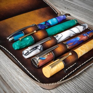 Puede incluir: Un estuche de cuero marr&oacute;n con cremallera y seis ranuras para bol&iacute;grafos. El estuche est&aacute; abierto y se pueden ver cinco bol&iacute;grafos. Los bol&iacute;grafos est&aacute;n hechos de diferentes materiales y tienen varios colores y patrones, incluyendo azul, verde, blanco y amarillo.