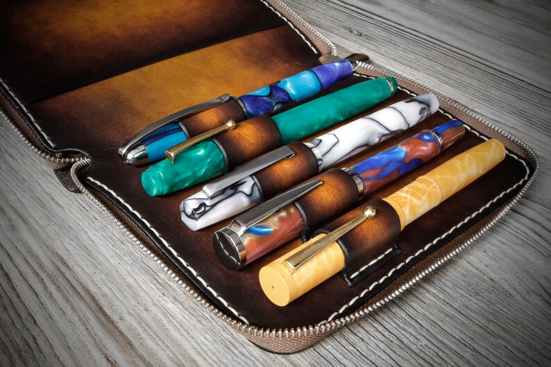 Puede incluir: Un estuche de cuero marr&oacute;n con cremallera y seis ranuras para bol&iacute;grafos. El estuche est&aacute; abierto y se pueden ver cinco bol&iacute;grafos. Los bol&iacute;grafos est&aacute;n hechos de diferentes materiales y tienen varios colores y patrones, incluyendo azul, verde, blanco y amarillo.