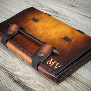 May include: A brown leather journal with a black pen holder and a black strap. The journal has a distressed look and is personalized with the initials "MV".