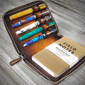 Puede incluir: Una funda de cuero marr&oacute;n con cremallera que tiene seis ranuras para bol&iacute;grafos y un bolsillo para una libreta. La funda est&aacute; abierta y muestra una pila de libretas Field Notes y varios bol&iacute;grafos con dise&ntilde;os coloridos.