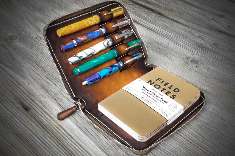 Puede incluir: Una funda de cuero marr&oacute;n con cremallera que tiene seis ranuras para bol&iacute;grafos y un bolsillo para una libreta. La funda est&aacute; abierta y muestra una pila de libretas Field Notes y varios bol&iacute;grafos con dise&ntilde;os coloridos.