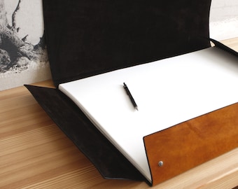 May include: A brown leather portfolio with a black leather flap and a white sheet of paper inside. A black pen is lying on the paper.