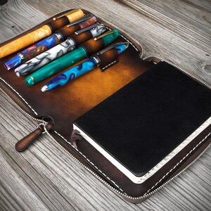Puede incluir: Un estuche de cuero marr&oacute;n con cremallera que contiene un cuaderno negro y seis bol&iacute;grafos. El estuche tiene un tirador de cremallera y costuras blancas.