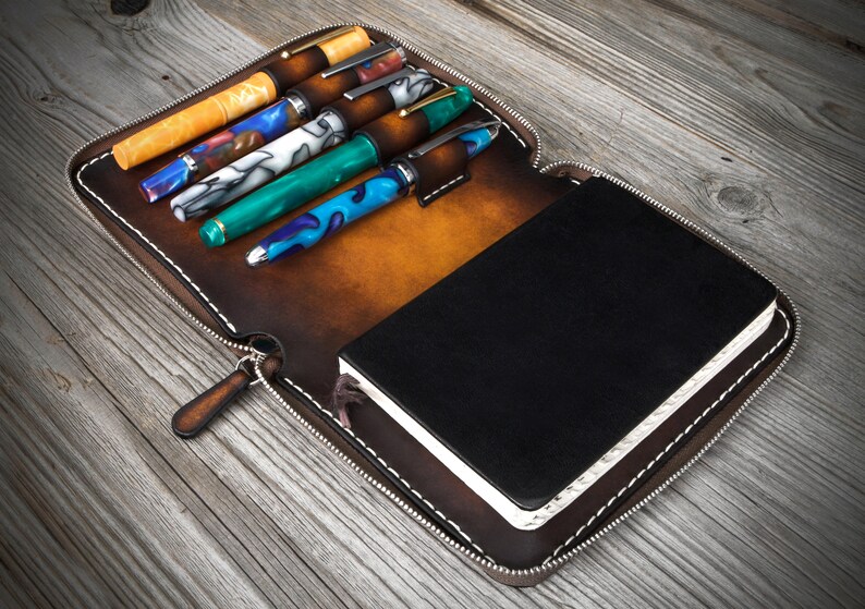 Puede incluir: Un estuche de cuero marr&oacute;n con cremallera que contiene un cuaderno negro y seis bol&iacute;grafos. El estuche tiene un tirador de cremallera y costuras blancas.