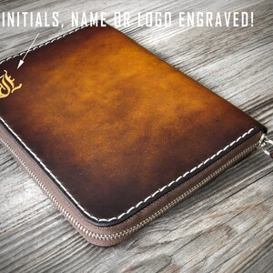 Puede incluir: Una cartera de cuero marr&oacute;n con cremallera y costuras blancas. La cartera tiene una inicial "J" grabada en oro en la parte delantera. El texto "YOUR INITIALS, NAME OR LOGO ENGRAVED!" es visible en la parte superior de la imagen.