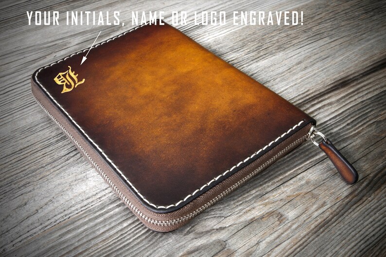 Puede incluir: Una cartera de cuero marr&oacute;n con cremallera y costuras blancas. La cartera tiene una inicial "J" grabada en oro en la parte delantera. El texto "YOUR INITIALS, NAME OR LOGO ENGRAVED!" es visible en la parte superior de la imagen.