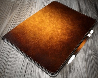May include: A brown leather tablet case with a matching stylus. The case has a textured surface and a stitched border. The stylus is brown with a white tip. The case and stylus are on a wooden surface.
