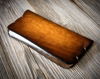 May include: A brown leather phone case with a worn, distressed look. The case is closed and lying on a wooden surface.
