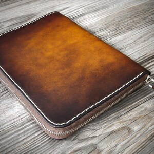 Puede incluir: Una cartera de cuero marr&oacute;n con cremallera y costuras blancas. La cartera est&aacute; cerrada y se encuentra sobre una superficie de madera.