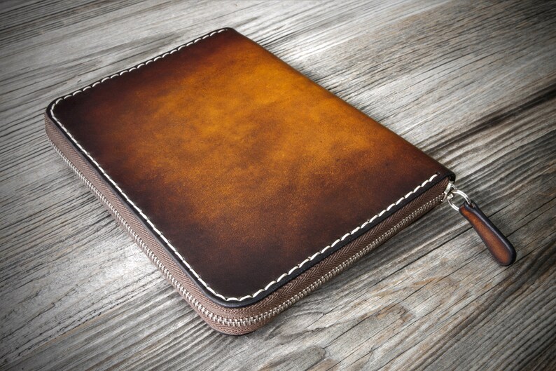 Puede incluir: Una cartera de cuero marr&oacute;n con cremallera y costuras blancas. La cartera est&aacute; cerrada y se encuentra sobre una superficie de madera.