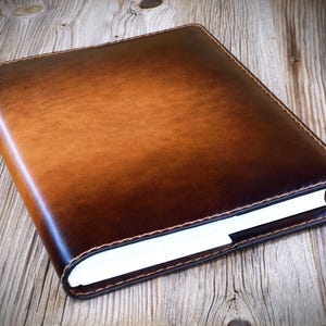 May include: A brown leather journal with a gradient effect, transitioning from light to dark. The journal is closed, revealing the white pages within. The edges are stitched with a contrasting thread, resting on a wooden surface.