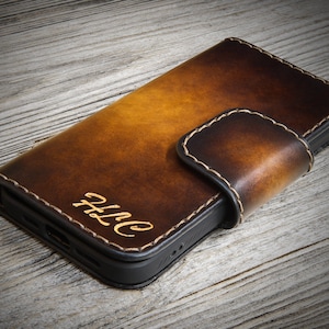 Peut inclure: Une coque de téléphone en cuir marron avec une fermeture à rabat et un monogramme "ALC" en or gaufré.
