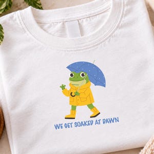 May include: A white t-shirt featuring a graphic of a green frog wearing a yellow raincoat and yellow boots while holding a blue polka-dot umbrella. Below the frog, the text reads "WE GET SOAKED AT DAWN" in blue capital letters.