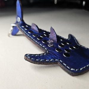 Może przedstawiać: Brelok do kluczy w kształcie rekina wielorybiego wykonany ze skóry w kolorze niebieskim, ze szwami w kolorze białym i srebrnymi kropkami. Brelok ma pętelkę do przypięcia do torby lub kluczy.