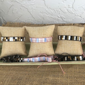 May include: Five colorful bracelets with different patterns and colors, displayed on small fabric cushions. The bracelets are made of small square tiles and have a variety of colors, including pink, white, black, and gold.