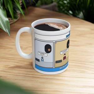 May include: A white ceramic mug featuring a two-panel comic strip. The first panel shows a robot saying, I CAN ANSWER ANY QUESTION. The second panel shows a person asking, WHAT'S FOR DINNER TOMORROW? The mug has a blue border and dark coffee inside.