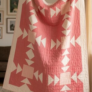 May include: A rectangular quilt featuring a geometric pattern of dusty rose and off-white fabric. The design consists of repeating squares and triangles arranged in a symmetrical layout with visible straight-line quilting stitches throughout.