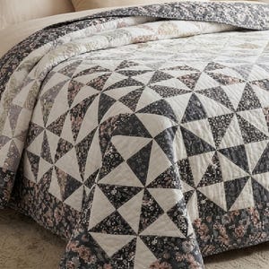 May include: A quilted bedspread featuring a geometric pattern of triangles arranged in a pinwheel design. The fabric displays a mix of dark charcoal gray, off-white, and muted dusty pink floral and paisley prints throughout the quilt.