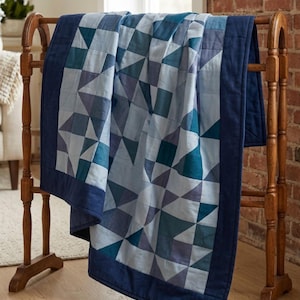 May include: A quilt featuring a geometric patchwork pattern in various shades of blue, including navy, teal, and light blue. The quilt is constructed from triangular and square fabric pieces and is finished with a solid navy blue border.