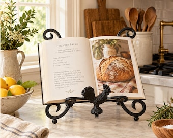 May include: A black metal cookbook stand featuring a decorative rooster figure at the base and scrolled accents. The stand holds an open book displaying a recipe for Country Bread with a photograph of a round loaf of bread on a cloth.