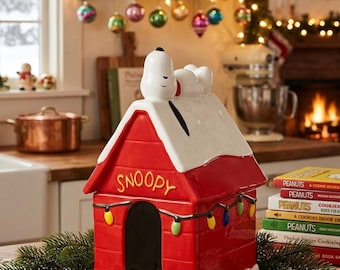 Könnte beinhalten: Ein Keramik-Keksglas in Form einer roten Hundehütte mit weißem Dach und weißem Sockel. Die weiße Snoopy-Figur liegt auf dem Dach. Die Vorderseite zeigt das Wort Snoopy in Gelb und eine Lichterkette mit bunten Weihnachtslichtern.