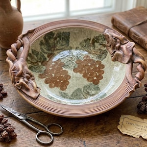 May include: A round ceramic bowl with a cream-colored interior featuring painted brown grape clusters and green leaves. The rim is textured with sculpted brown vine and leaf motifs. A small paper tag reads Terrafirma Pottery New York.
