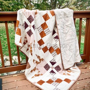 Puede incluir: Una colcha de patchwork con un patrón geométrico de cuadrados de tela en blanco, naranja quemado y granate oscuro. El diseño incluye colores sólidos y estampados florales, con un borde marrón y un reverso de color crema sólido.
