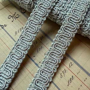 May include: Close-up of a roll of silver-gray decorative trim with a wavy, looped design. The trim is made of a woven material with a metallic sheen. The background is a vintage ledger paper with handwritten notes and numbers.