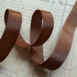 May include: A spool of brown grosgrain ribbon, with a ribbed texture, is curled on a vintage ledger. The ribbon's rich, warm color contrasts with the aged paper, creating a visual appeal. Ideal for crafts, sewing, and embellishments.