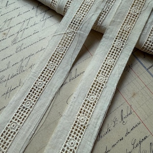 May include: Close-up of antique beige lace ribbon with a delicate openwork design. The ribbon features a repeating pattern of small circles within a grid, creating a textured effect. The ribbon is displayed on a vintage paper background with handwritten script.