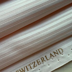 Puede incluir: Primer plano de una tela rosa pálido con rayas blancas verticales. La tela es texturizada y parece ser un material tejido. Las palabras "IN SWITZERLAND" están impresas en oro.