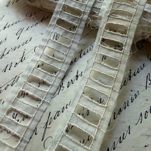 May include: Close-up of cream-colored, openwork ribbon. The ribbon features a series of rectangular cutouts, creating a delicate, lace-like effect. The ribbon is slightly crumpled, suggesting a vintage or antique aesthetic. The background is a handwritten script.
