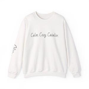 May include: A white long-sleeve crewneck sweatshirt featuring the black text "Calm. Cozy. Cavalier." printed across the chest. The right sleeve displays a small black graphic of a dog face with floppy ears and a simple line drawing.
