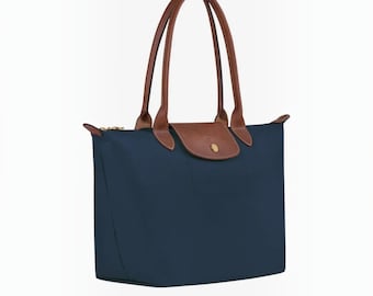 May include: A navy blue tote bag featuring brown leather handles and a matching brown leather flap closure with a gold-tone snap button. The bag has a structured trapezoidal shape with a zipper top and clean, minimalist lines throughout.